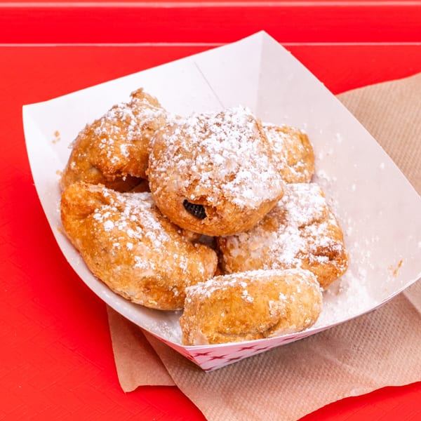 Deep-fried apple pies dusted with powdered sugar in a white paper tray on a red surface.