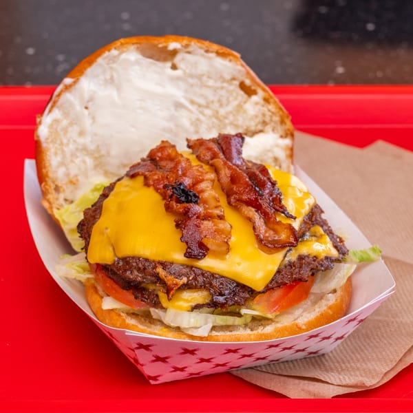 A Chili Hots double bacon cheeseburger with lettuce and tomato in a paper tray on a red surface.