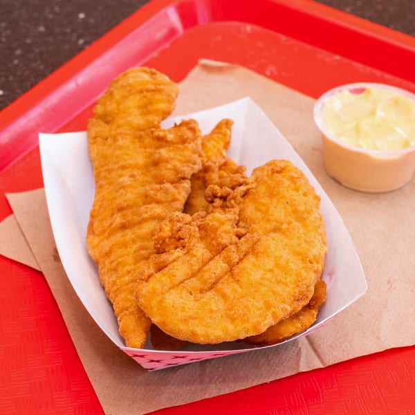 Crispy fried chicken tenders in a paper tray with a side of dipping sauce on a red tray.