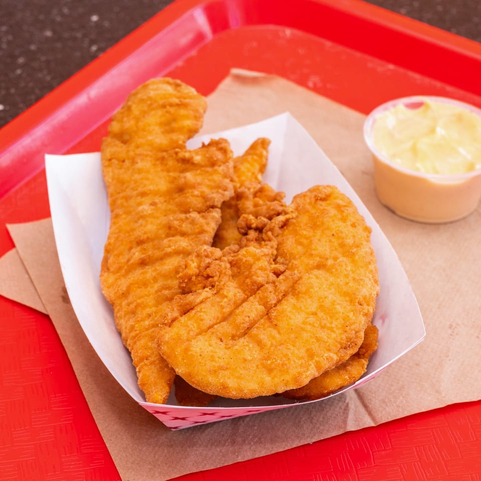 Crispy fried chicken tenders in a paper tray with a side of dipping sauce on a red tray.